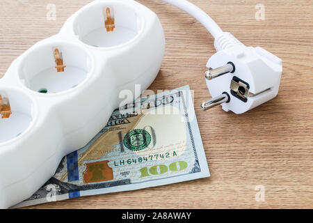 White electrical power strip and power plug on a one hundred dollar bill over brown wooden surface. Cost of electricity and expensive energy concepts. Stock Photo