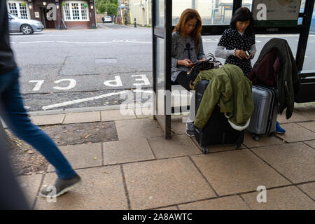 two asian women sitting at a bus stop with suitcases while people walk by Stock Photo