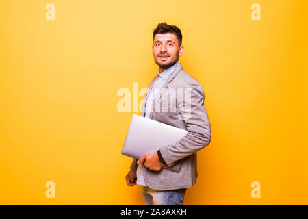 Thoughtful man with a beard dressed in a white T-shirt stands on a yellow background, keeps the laptop in his hands, looks at the screen and thinks.Ma Stock Photo