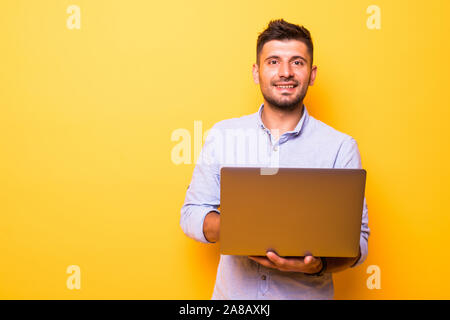Thoughtful man with a beard dressed in a white T-shirt stands on a yellow background, keeps the laptop in his hands, looks at the screen and thinks.Ma Stock Photo