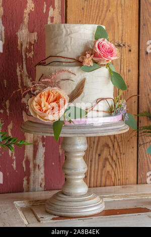 A rustic wedding cake on a stand with flowers and sticks wrapping the cake. The cake sits on top of a door with barn wood behind. Stock Photo