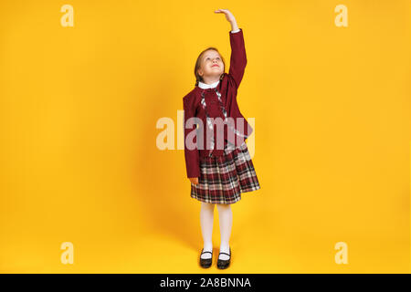 A full-length schoolgirl child in uniform is measuring her height. Little girl on a yellow background. Stock Photo