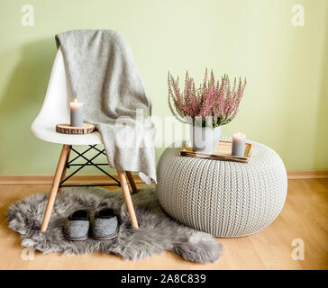 Plastic chair with wood legs covered with gray wool plaid, knitted pattern plastic table, fake sheepskin rug with felt slippers and heather in flower Stock Photo