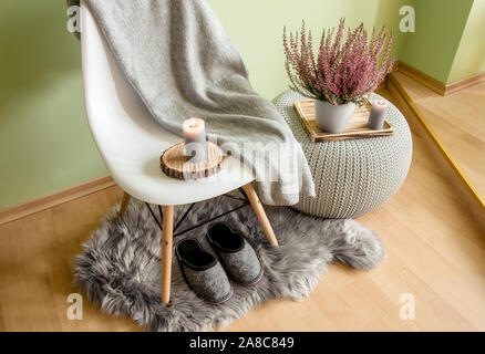 Plastic chair with wood legs covered with gray wool plaid, knitted pattern plastic table, fake sheepskin rug with felt slippers and heather in flower