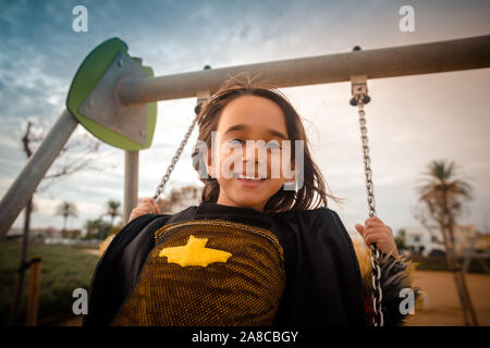 Close-up shot of a young European girl happy playing on a swing at an outdoor park wearing a bat costume. Stock Photo