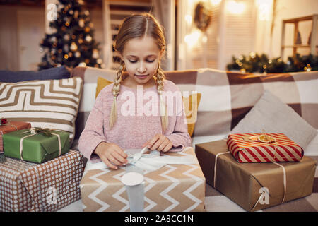 Little girl sitting on sofa and opening big Christmas present she getting it from Santa Claus for Christmas at home Stock Photo