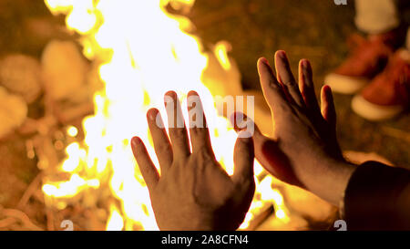 Close up of tourist sitting near the camp fire at night with his friends. Camper warming up his hands. Handheld footage Stock Photo