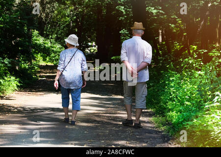 Middle aged couple is walking in forest. Woods on sides of trail. Elderly couple is walking in green park on holiday. Sunny summer day. Stock Photo