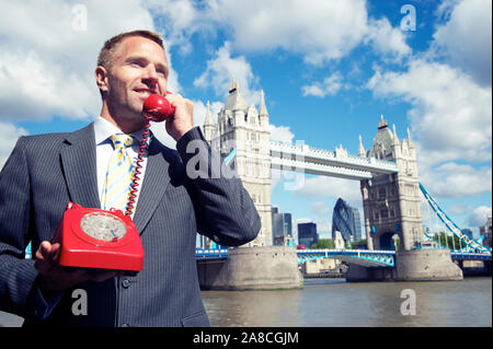 British businessman listening to the good new hotline on a old red rotary telephone in front of the London skyline at Tower Bridge Stock Photo