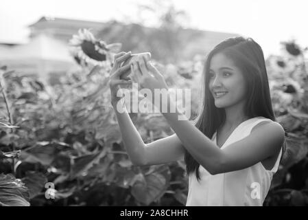 Young happy Asian woman smiling while taking selfie picture with mobile phone in the field of blooming sunflowers Stock Photo