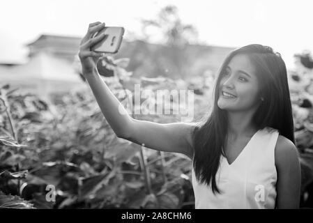 Young happy Asian woman smiling while taking selfie picture with mobile phone in the field of blooming sunflowers Stock Photo