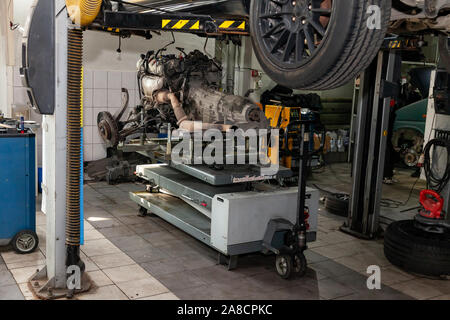 A used car raised on a lift for repair and under it a detached engine and gearbox suspended on a gray lift table near workbench in a vehicle repair sh Stock Photo