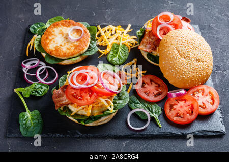 juicy turkey burgers on buns with shredded marble cheese, crispy fried bacon, tomato slices, spinach leaves, and red onion on a black slate plate, vie Stock Photo