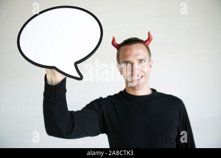 Smiling man with red devil horns holding a large cartoonish speech bubble looking at the camera with a mischievous expression Stock Photo