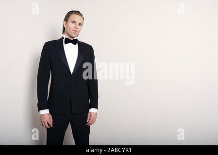 Young man with long hair standing in full tuxedo against plain white wall looking at the camera with a serious expression Stock Photo