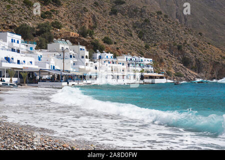 Loutro, Chania, Crete, Greece. Waves breaking on stony beach, waterfront tavernas in background. Stock Photo