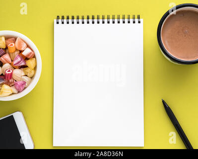 Open notepad on spiral with clean white page, cup with cocoa, caramels in a bowl, smartphone and pencil on a bright yellow background color Stock Photo