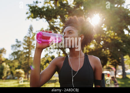 Fitness athlete young african american woman listening to music on earphones drinking water in a reusable water bottle after working out exercising on Stock Photo