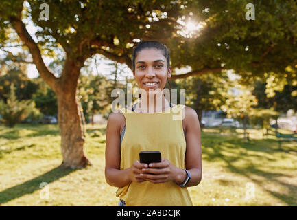 Portrait of a smiling young sporty woman holding mobile phone in hand looking at camera in the park - fit woman smiling at the camera in a park on a Stock Photo