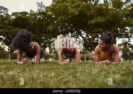 Three sporty young women smiling while doing plank exercises lying on green grass in the park Stock Photo