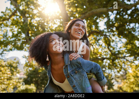 Two diverse multiracial friends having fun laughing and giving a piggyback to each other in the park on a warm summer day