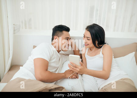 Young asian couple smile listen music with smart mobile phone on bed at bedroom with fun and enjoy Stock Photo