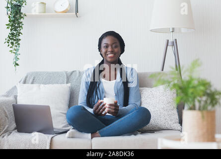 Afro woman freelancer sitting on couch with coffee and laptop Stock Photo