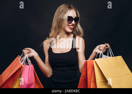 Young pretty girl in sunglasses posing with a bunch of coloured paper bags isolated black background Stock Photo