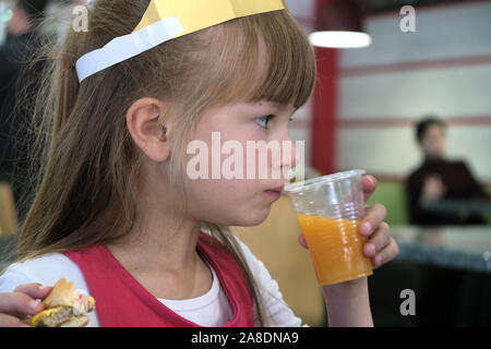 Close up of cute child girl drinking orange juice from plastic cap and eating fast food in a restaurant. Stock Photo