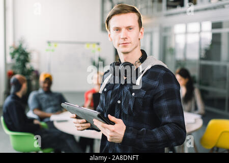 Half-length portrait of cheerful young manager or designer dressed in casual outfit and earphones, having work break in office, holding digital tablet Stock Photo