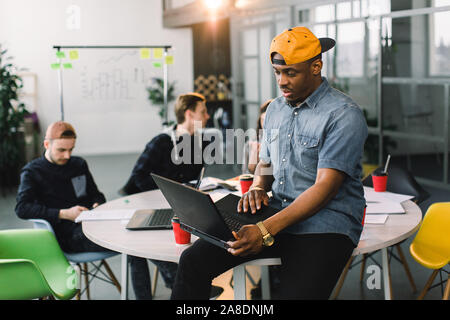 Dark skinned African american coworker in jeans shirt and yellow cap and using laptop in modern office. Multiracial young creative people are working Stock Photo