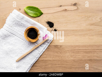 Raw activated charcoal powder in wooden cup and natural bamboo toothbrush next to it, green leaf and bath towel. Charcoal toothpaste concept. Lot of c Stock Photo