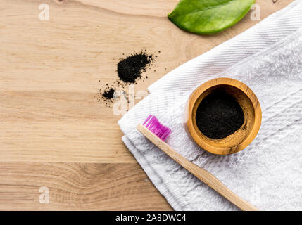 Raw activated charcoal powder in wooden cup and natural bamboo toothbrush next to it, green leaf and bath towel. Charcoal toothpaste concept. Lot of c Stock Photo