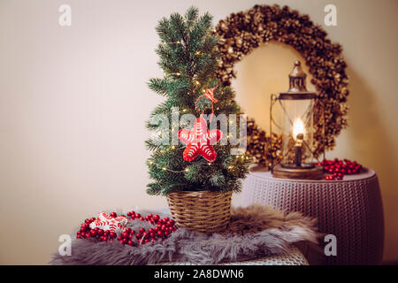 Small portable tabletop decorated Christmas tree on table, party lights illuminated, red felt fabric ornament. Light bulb lantern with pine cone wreat Stock Photo