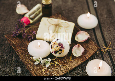 Various spa bath products on brown wooden tray creamy bath bomb, natural soap bar with cotton string, dried flower petals, spa candles lit, essential Stock Photo