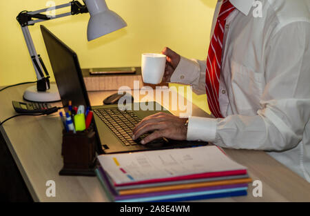 Man in suit works with laptop in office and drinks coffee. Businessman in white shirt with red tie works Stock Photo