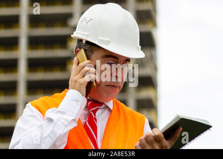 Construction engineer man in shirt and tie with safety helmet and vest works at construction site, talking phone. Concept of people working in industr Stock Photo