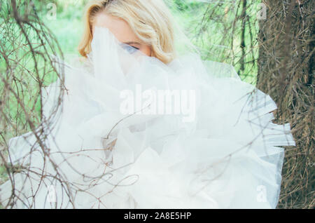 Portrait of young blond bride in forest through branches and veil, looking to the camera Stock Photo