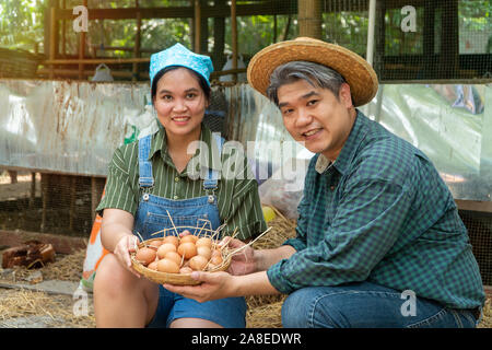 Asian couple farmer holding fresh chicken eggs into basket was sitting near hen beside chicken farm.Smiling because happy with the products from the f