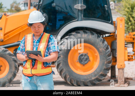 Woman Engineering wearing a white safety helmet standing In front of the backhoe And are using tablet to check the blueprint with construction Stock Photo