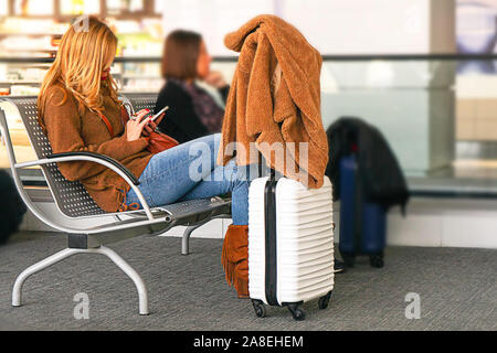White color suitcase and beautiful woman looking at cellphone, in airport departure lounge. Vacation concept, traveler layout. Selective focus. Stock Photo