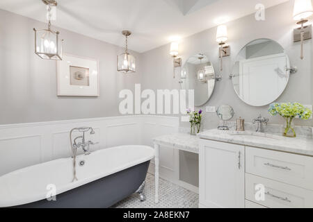 A beautiful white bathroom with a stand alone clawfoot bathtub with chrome hardware. Flowers are on the white marble vanity counter top. Stock Photo