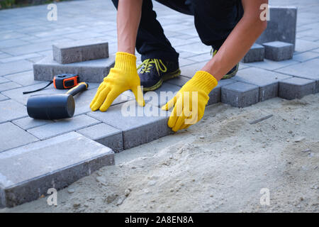 The master in yellow gloves lays paving stones in layers. Garden brick pathway paving by professional paver worker. Laying gray concrete paving slabs Stock Photo