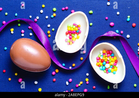 Chocolate egg with ribbon and colored candies on blue background. Easter background presentation with place for text. Stock Photo