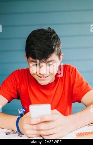 Handsome Caucasian 13-year old boy wearing red t-shirt sitting outdoors using electronic gadget. Schoolboy holding mobile phone, messaging friends Stock Photo