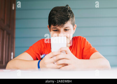Handsome Caucasian 13-year old boy wearing red t-shirt sitting outdoors using electronic gadget. Schoolboy holding mobile phone, messaging friends Stock Photo