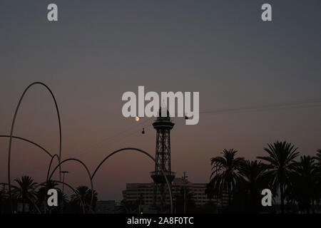 Silhouette of Barcelona cable car tower, palm trees, the moon & structures at sunset Stock Photo