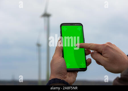 man using his smartphone in front of out of focus wind turbines. Accessing iot wind energy network with a cell phone. Using mobile phone outdoors Stock Photo