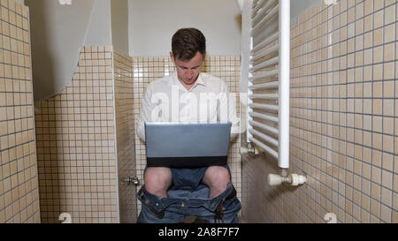 Inspired man workaholic is working in toilet on computer. He is typing something very quickly and thinking about it. Quiet place to work. Stock Photo