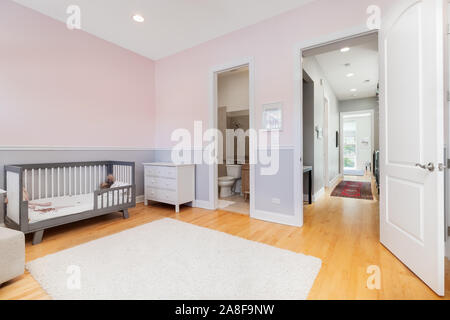 A children's bedroom with white and purple walls looking down the hallway. A crib sits up against the wall and a rug over the light hardwood floors. Stock Photo